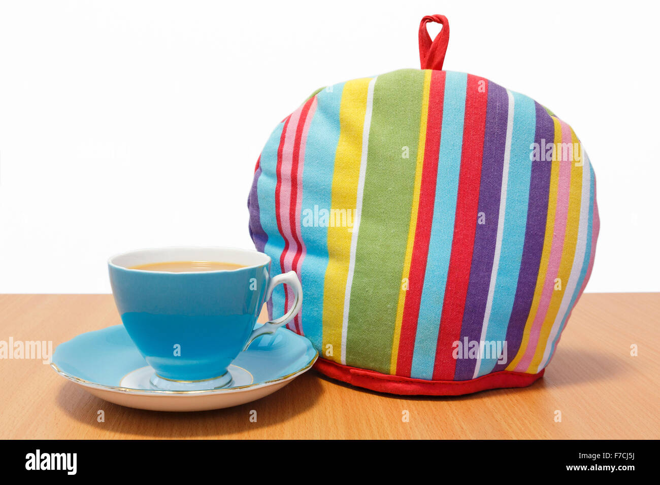 Bone china cup of tea with a saucer beside a colourful striped teacosy covering a teapot to keep it warm on a wooden tabletop. Stock Photo