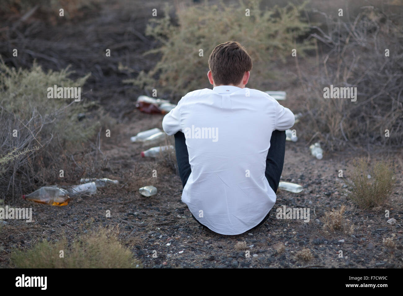Teenage boy drinking alcohol outside, sitting in the dirt with empty bottles. Stock Photo