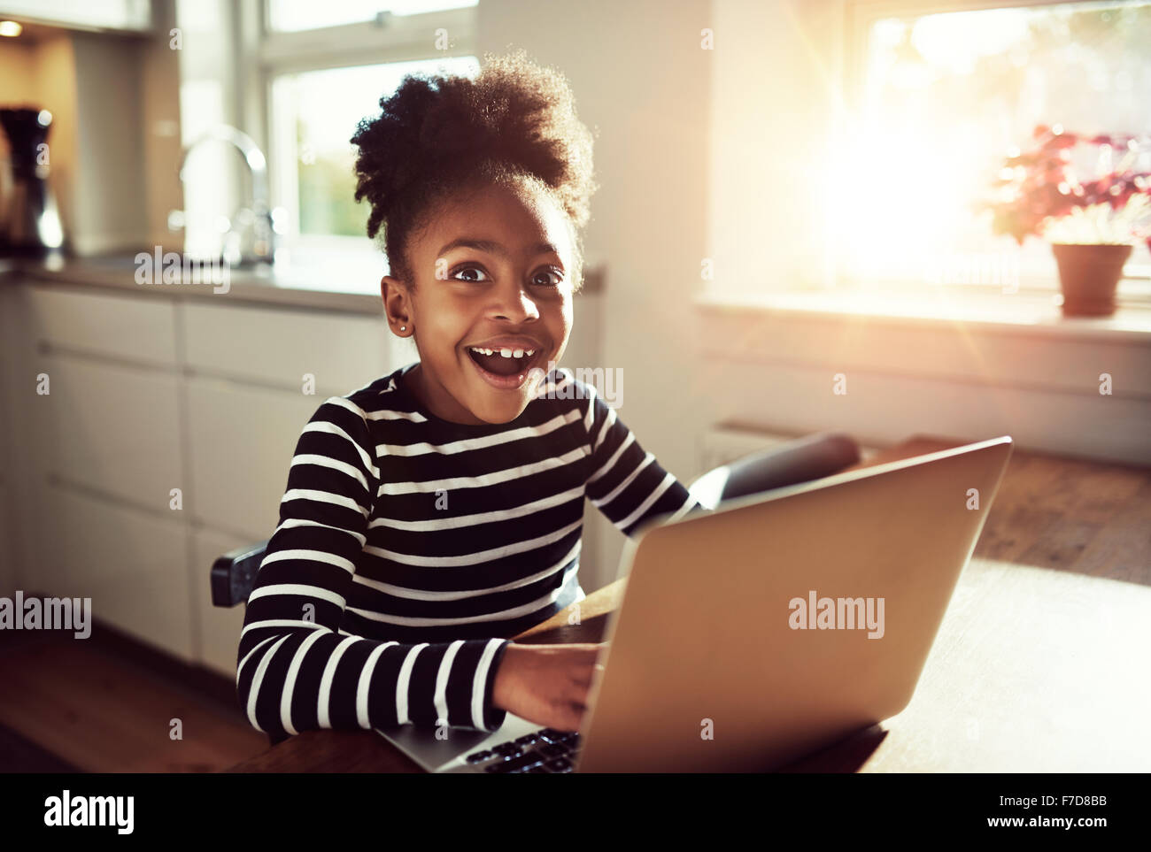 Black girl sitting playing on a laptop computer at home looking at the camera with a joyful expression of amazement and wonder Stock Photo