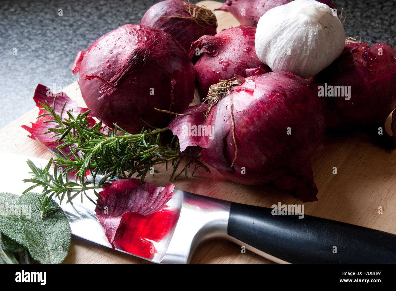 Whole red onions, a garlic bulb, rosemary and sage on a wooden chopping board with a chefs knife Stock Photo