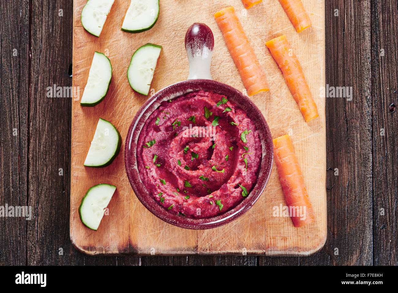 Hummus with beetroot in bowl on cutting board, surrounded by carrots and cucumber, on a dark wood table Stock Photo
