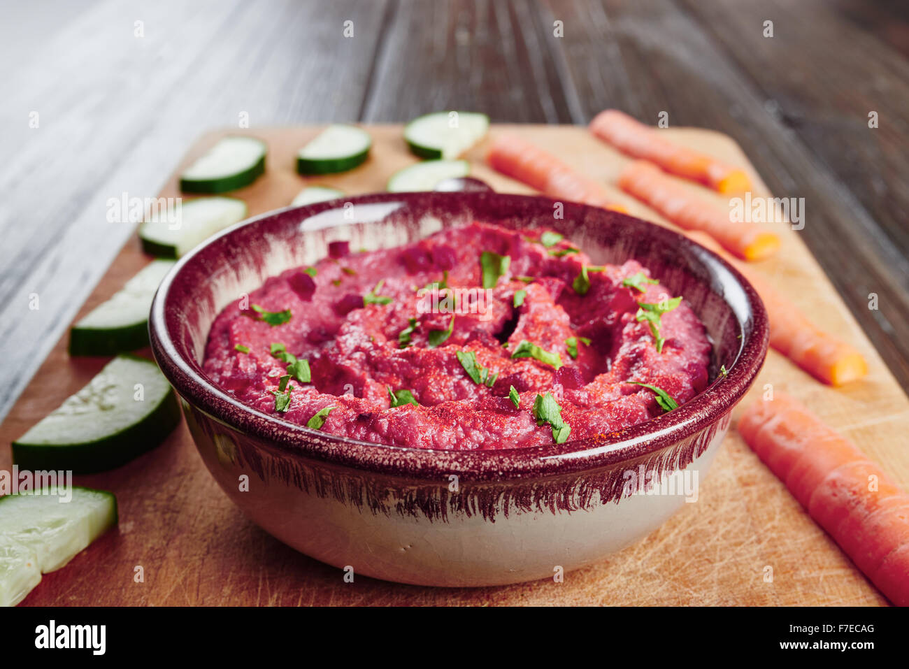 Hummus with beetroot in bowl on cutting board, surrounded by carrots and cucumber, on a dark wood table Stock Photo