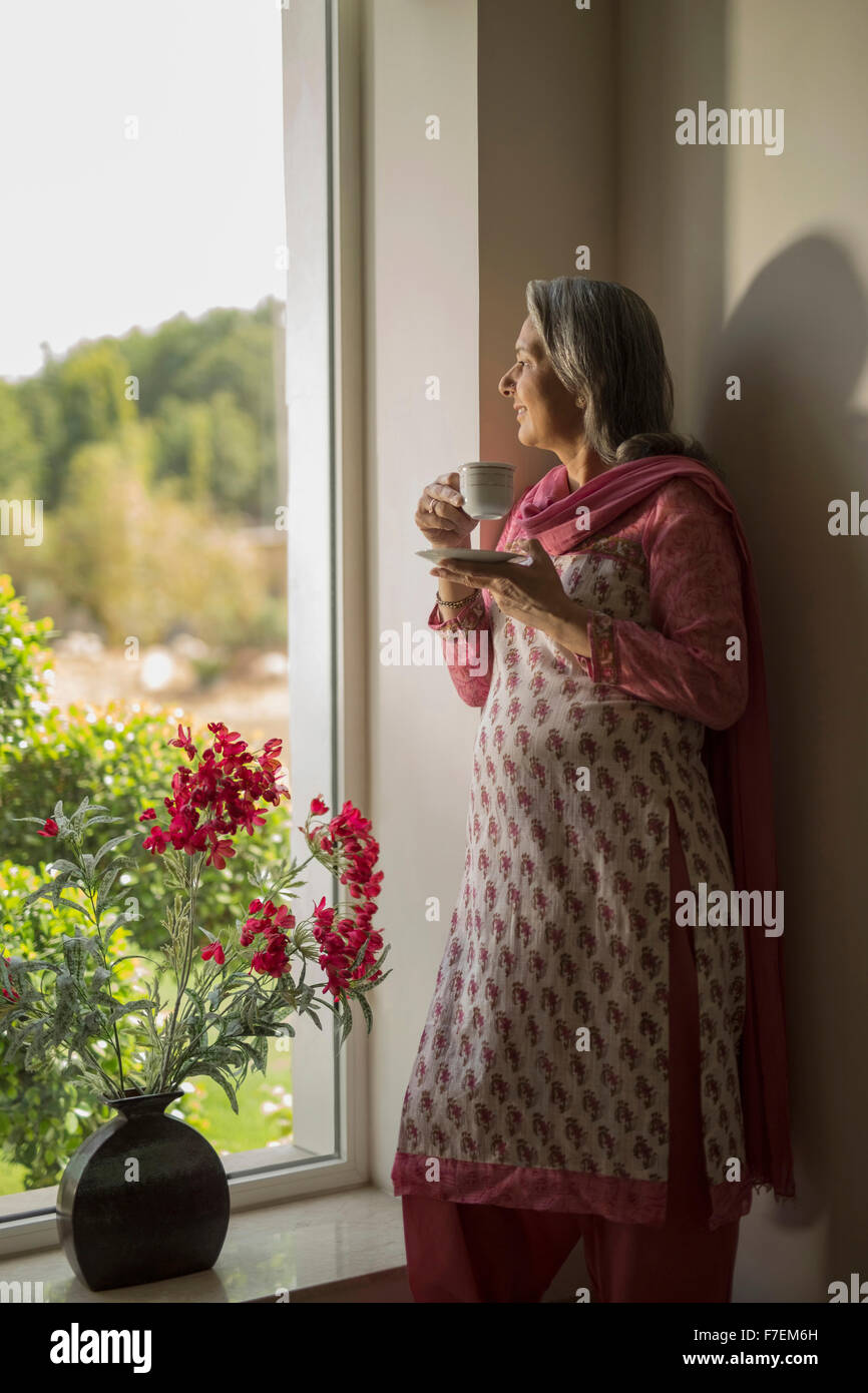 Older woman standing at window with cup of tea Stock Photo