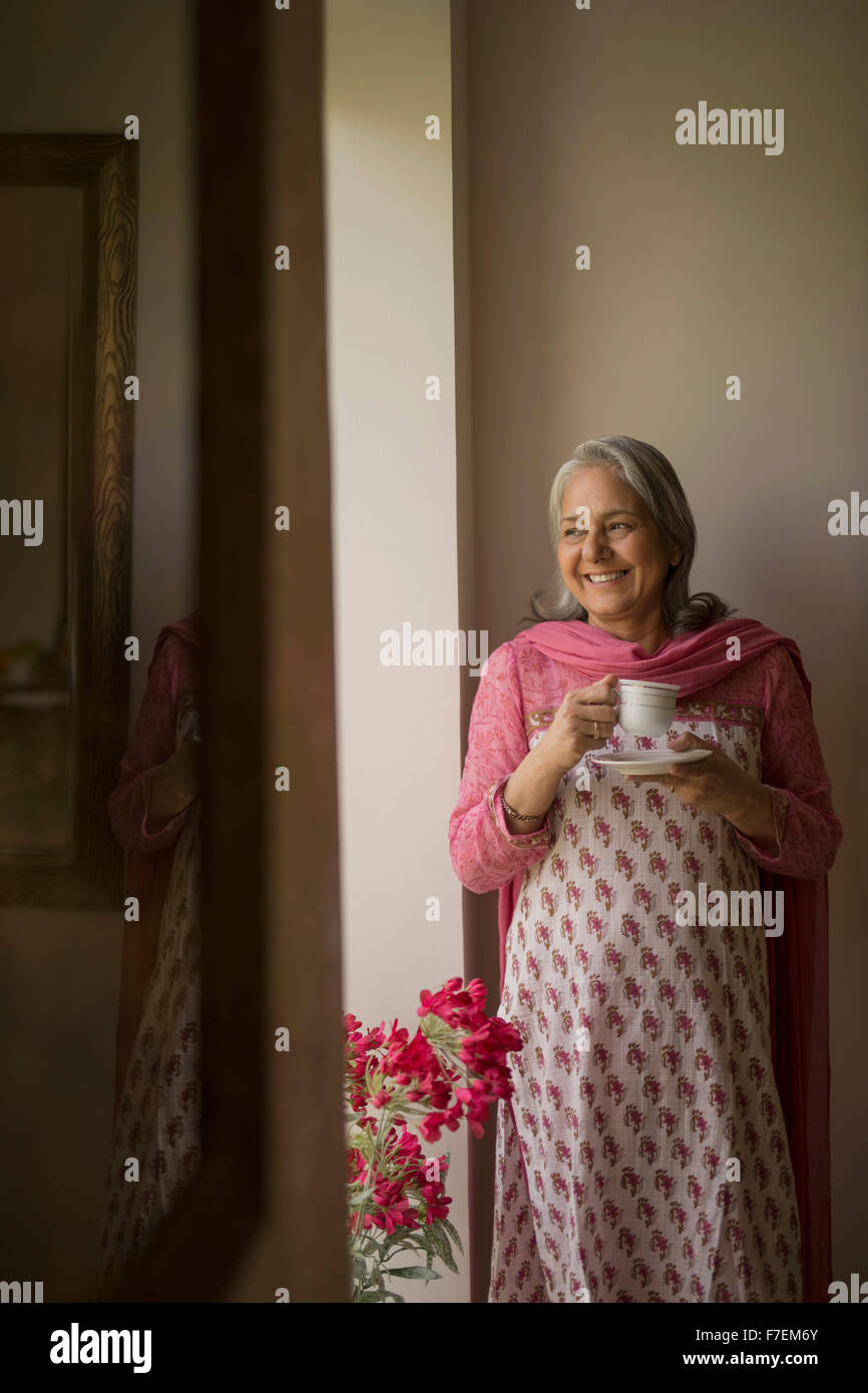Mature woman standing at window with tea cup smiling Stock Photo