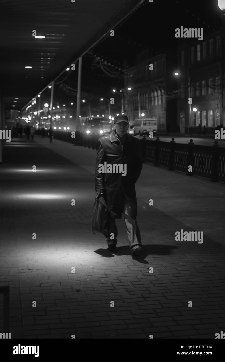 An elderly gentlemen walks through a pool of light on a pedestrian footpath in a busy city at night Photograph is in monochrome Stock Photo