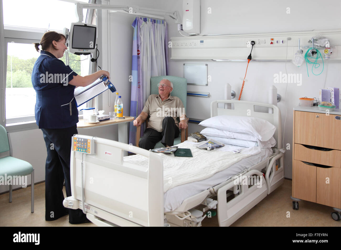 A nurse talks to an elderly patient in a modern UK hospital ward. Both are smiling and looking happy. (Names and medical data removed for anonymity) Stock Photo