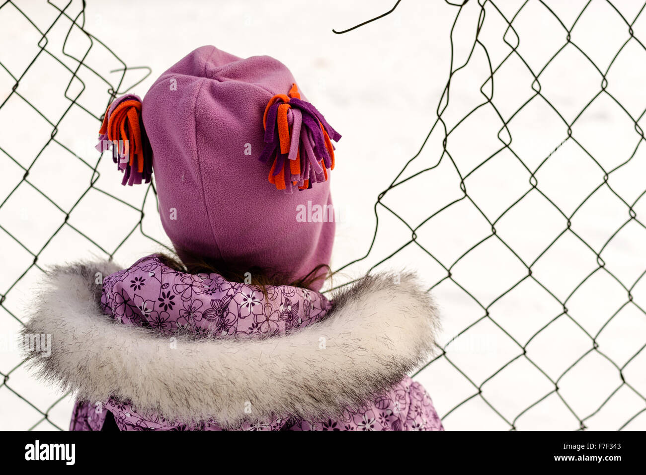 Young girl in warm winter clothes looks through a broken chain link fence Stock Photo