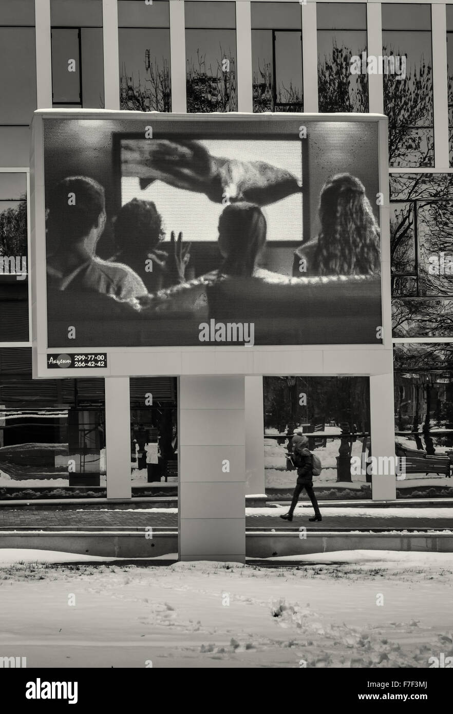Girl on her smartphone walks underneath a huge outdoor advertising Television showing a happy nuclear family watching television Stock Photo