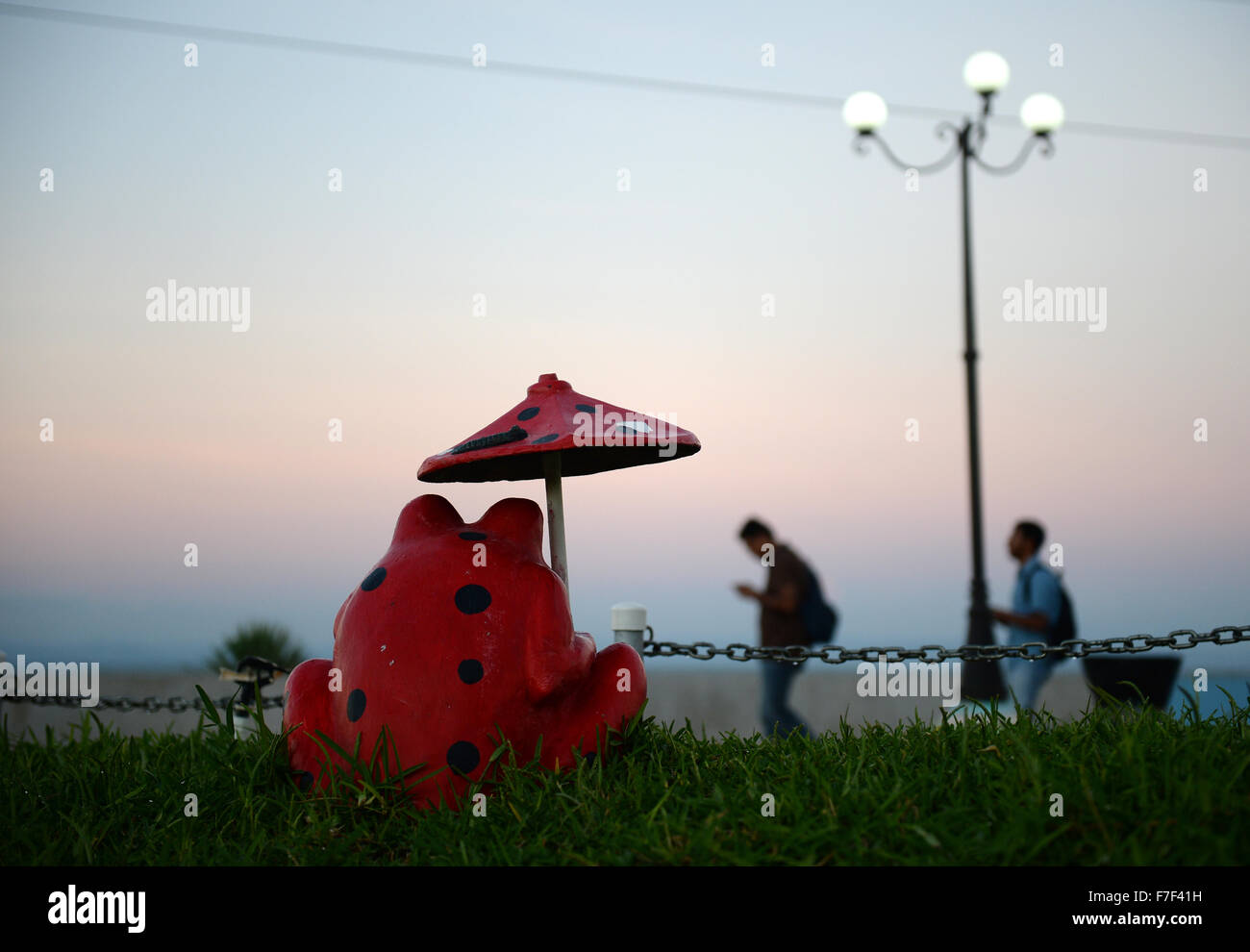 Managua, Nicaragua. 27th Nov, 2015. Strollers walk past a sculpture depicting a red frog with umbrella standing on the promenade of El Malecon at the Port Salvador Allende in Managua, Nicaragua, 27 November 2015. Photo: Jens Kalaene/dpa/Alamy Live News Stock Photo