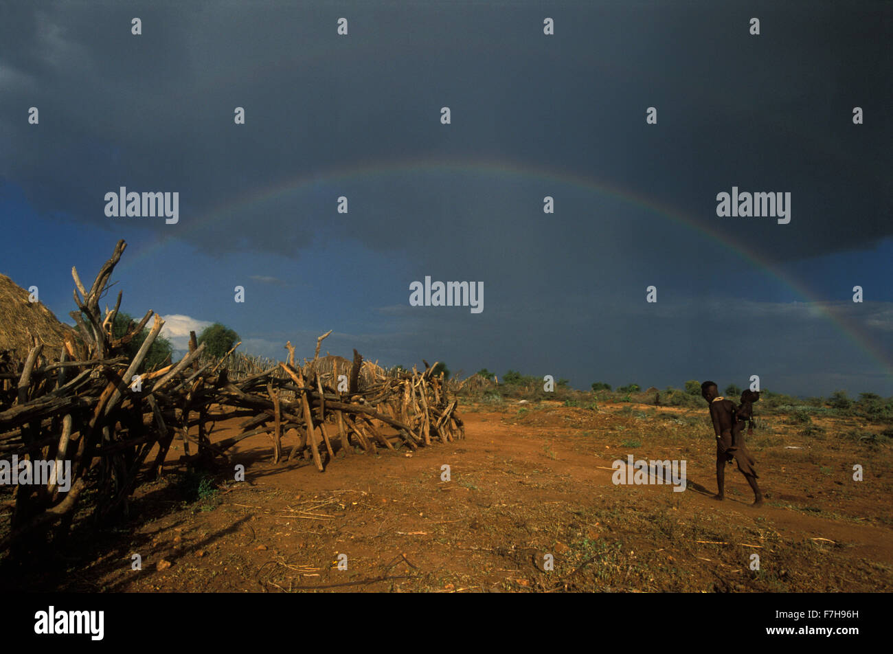 A double rainbow arches over a Hamar village and a Hamar girl who carries and infant on her hip, in South Omo, Ethiopia Stock Photo
