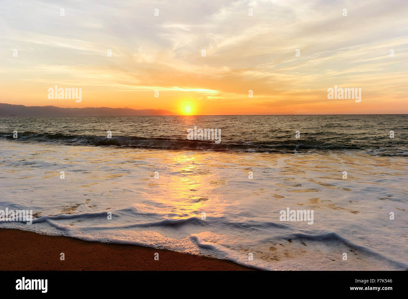 Beach sunset is a beautiful ocean sunset with a soft wave rolling toward the sand and the orange sun setting in the cloud filled Stock Photo