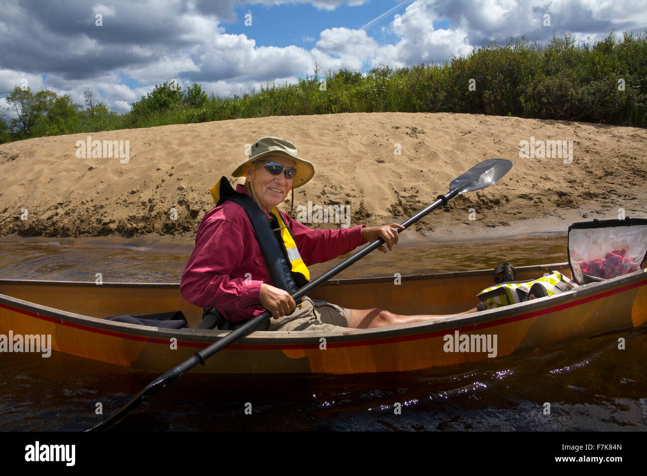 Senior man paddling a small canoe on Limekiln Lake in Old Forge, New York, with a kayak paddle. A sandy beach is in back. Stock Photo