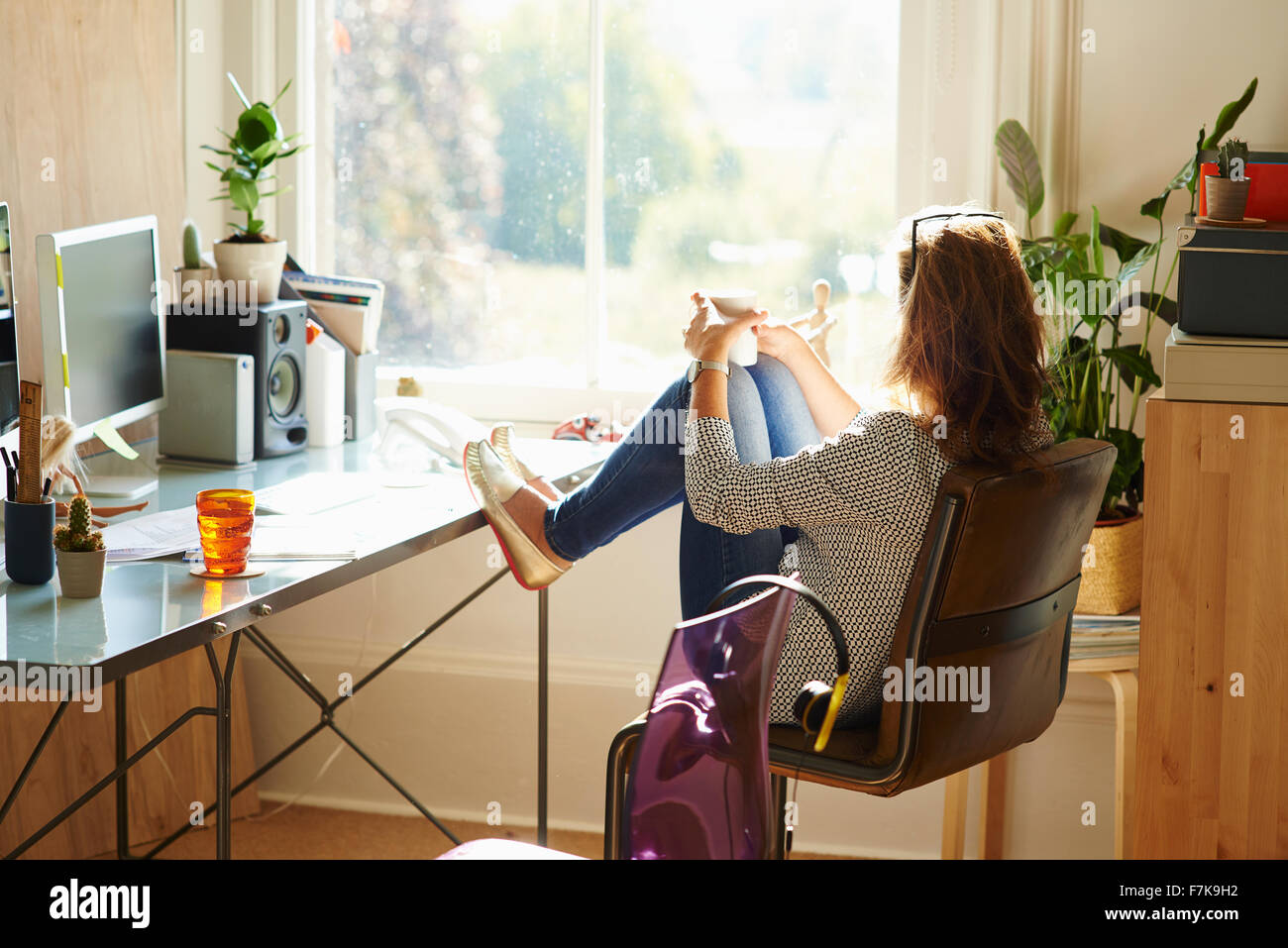 Pensive woman looking through window with feet up on desk in sunny home office Stock Photo