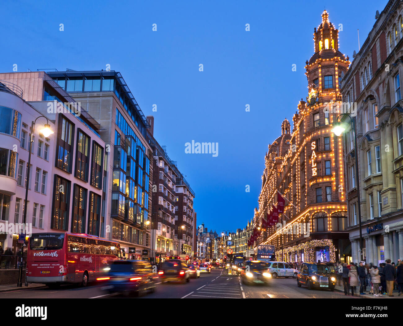 Knightsbridge Brompton Road shops including Harrods store at dusk with lit 'Sale' sign shoppers red buses and taxis Knightsbridge London SW1 Stock Photo