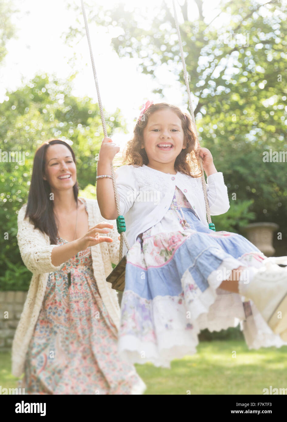 Mother in dress pushing daughter on swing in backyard Stock Photo