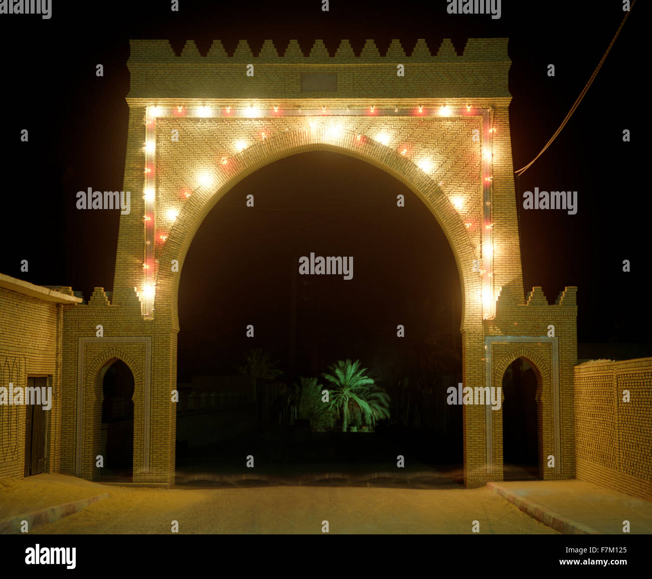 A lighted gate at night leads to the main Palmeraie in Tozeur. A palm tree is illuminated in the distance. Tozeur, Tunisia. Stock Photo