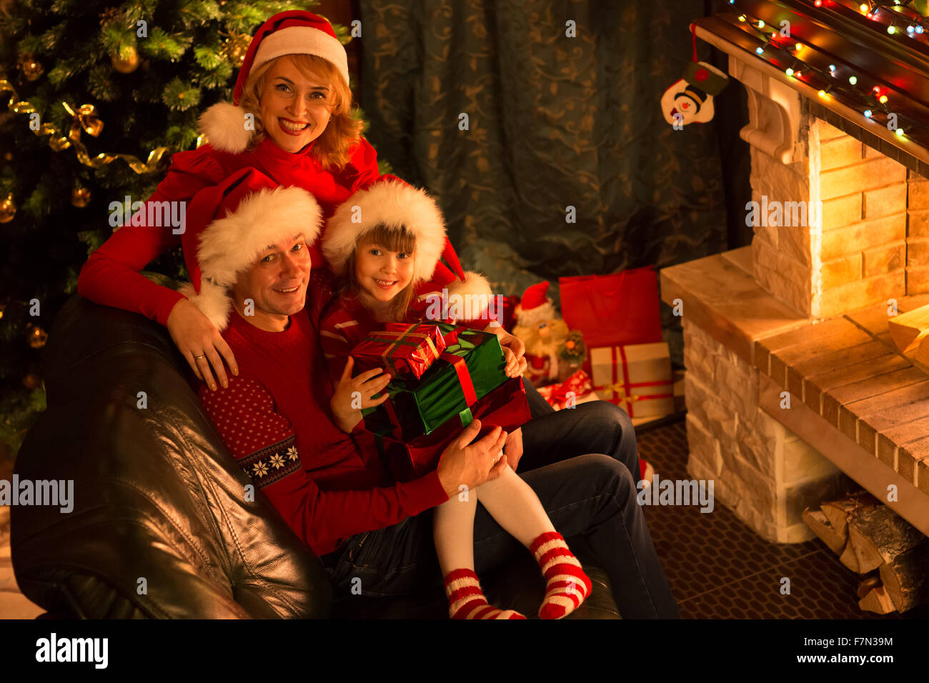 Happy family in red hats with gifts sitting at Christmas tree near fireplace Stock Photo