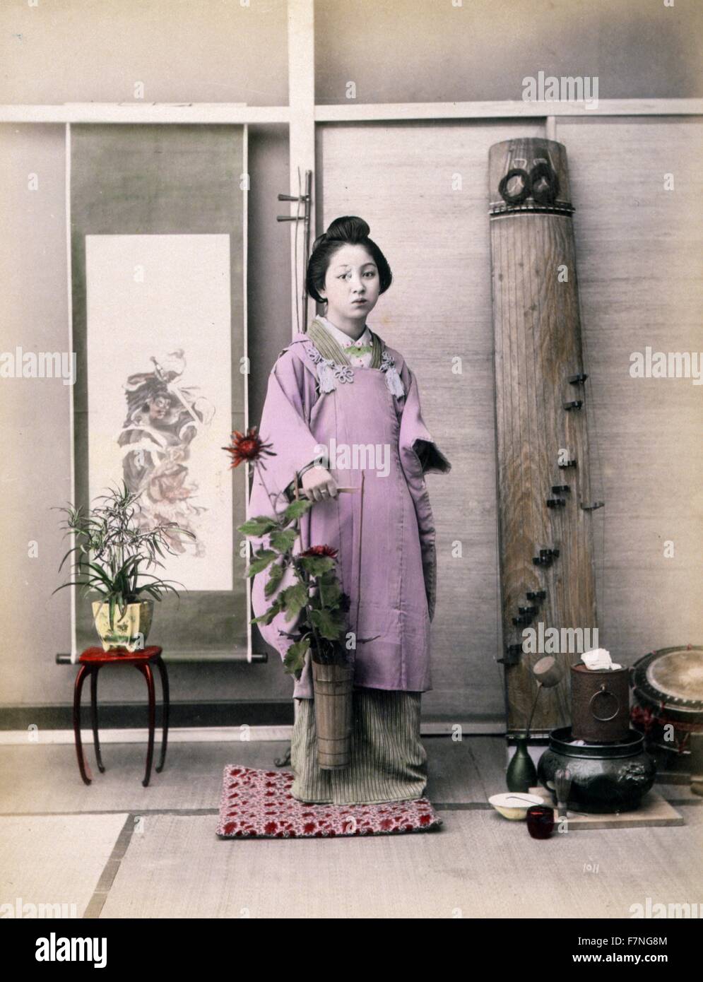 Photograph shows a portrait of a young woman, full-length, standing, facing front, holding a small, long-handled bucket with flowers in it. There is a small table with potted plant in front of a hanging scroll in the background on the left, and a Japanese zither leaning against the wall on the right. Watercolour drawings on the mount depict a Japanese red-crowned crane in flight with Mount Fuji in the background in the upper right and two cranes in the lower left. Stock Photo