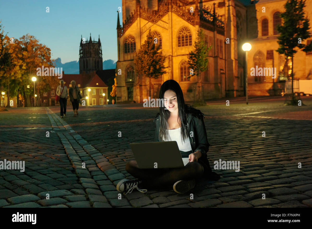Germany, Muenster, young woman with laptop sitting on pavement in the city at evening Stock Photo