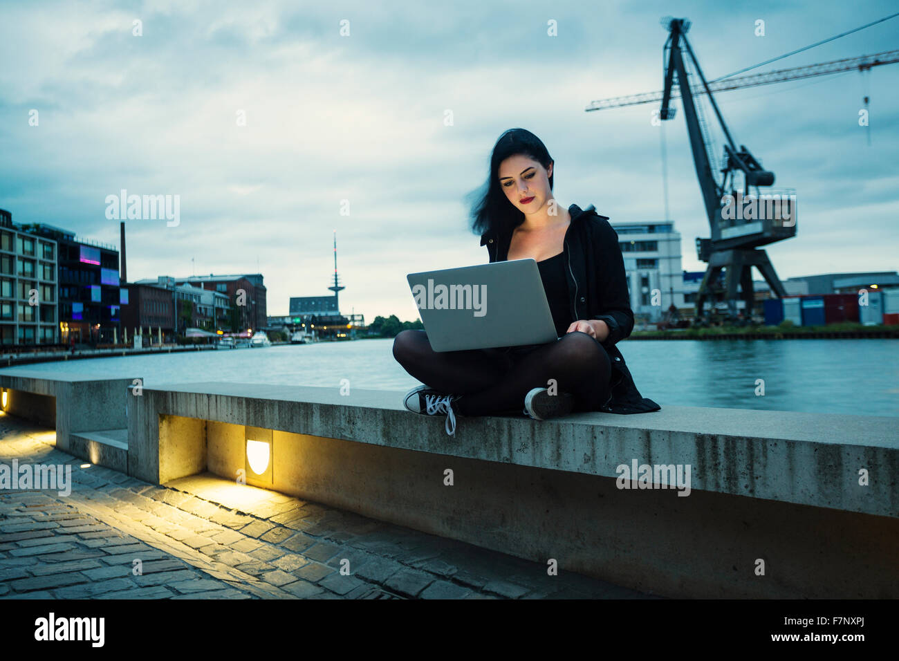 Germany, Muenster, young woman with laptop sitting in front of city harbour Stock Photo