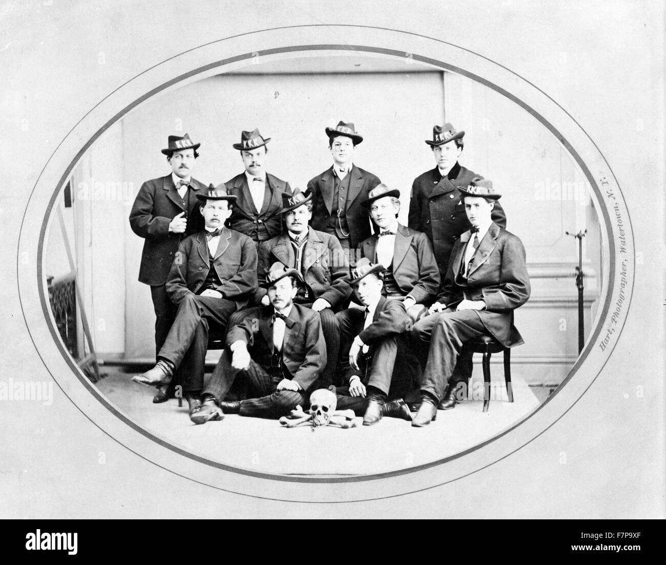 Photograph shows ten men posed, seated and standing, wearing hats with 'KKK' in large letters, and with a skull and bones arranged on the floor in front of them. Ku Klux Klan, Watertown Division, photographer, Watertown, New York. Stock Photo