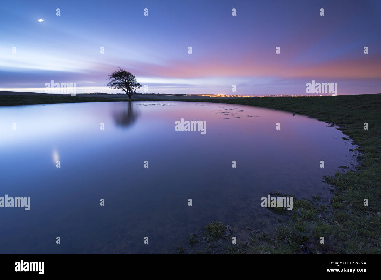 Dew pond near Ditchling Beacon, East Sussex, at night, with the moon reflected in the water, and city lights glowing on the horizon. Stock Photo