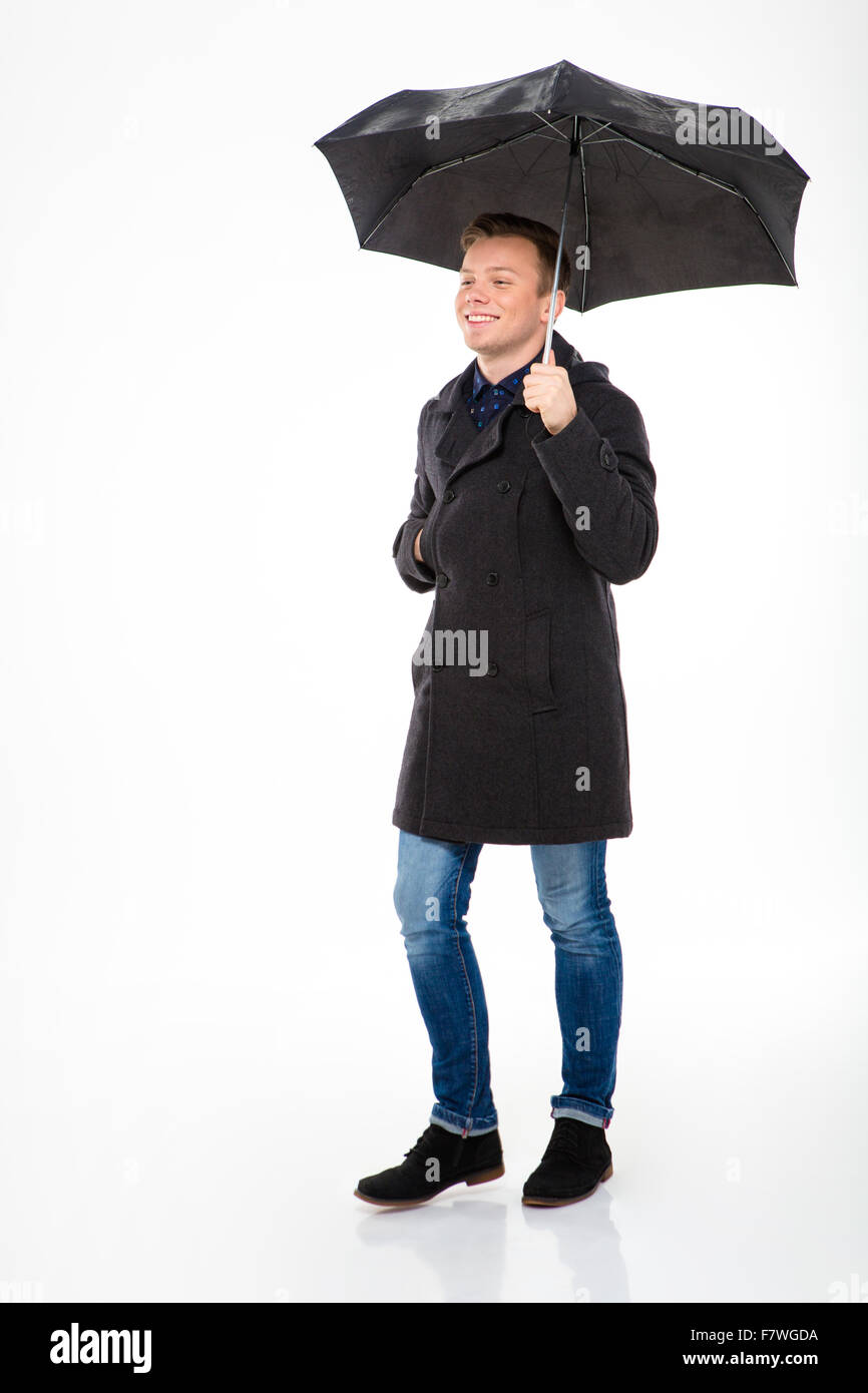 Happy content young male in coat and jeans walking under umbrella isolated over white background Stock Photo