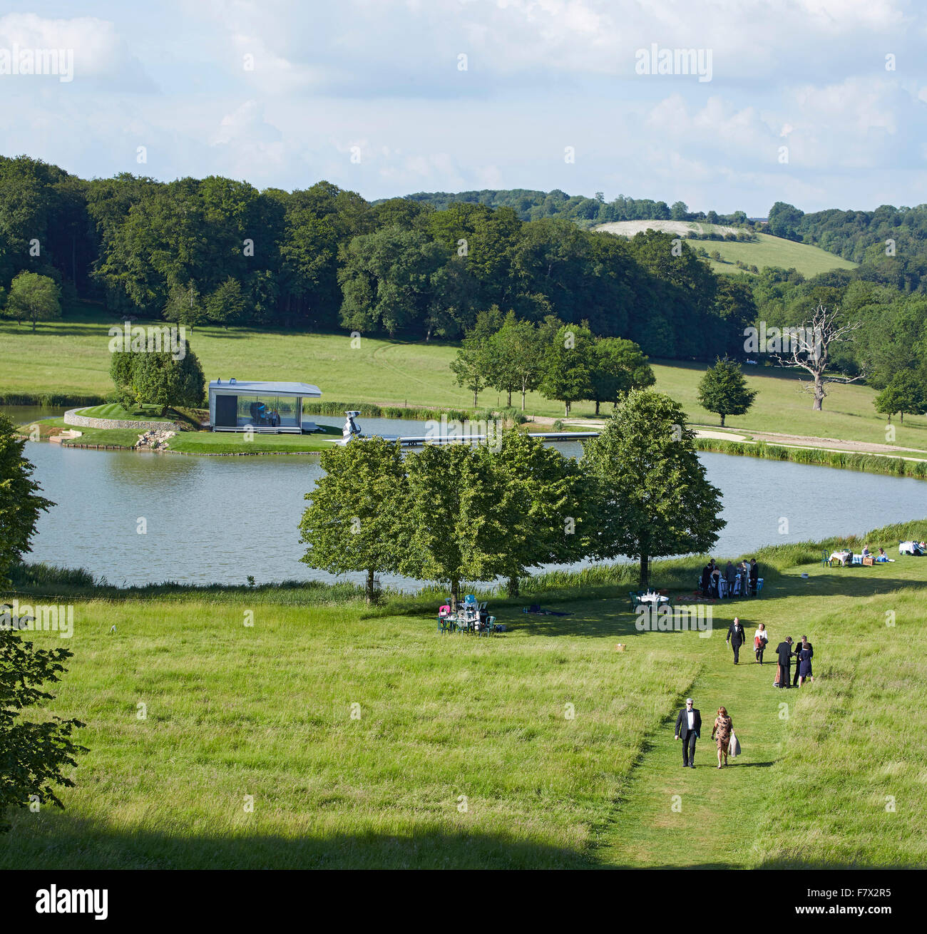 Distant elevation of glass pavilion in park with visitors. Island Pavilion and Footbridge, High Wycombe, United Kingdom. Archite Stock Photo