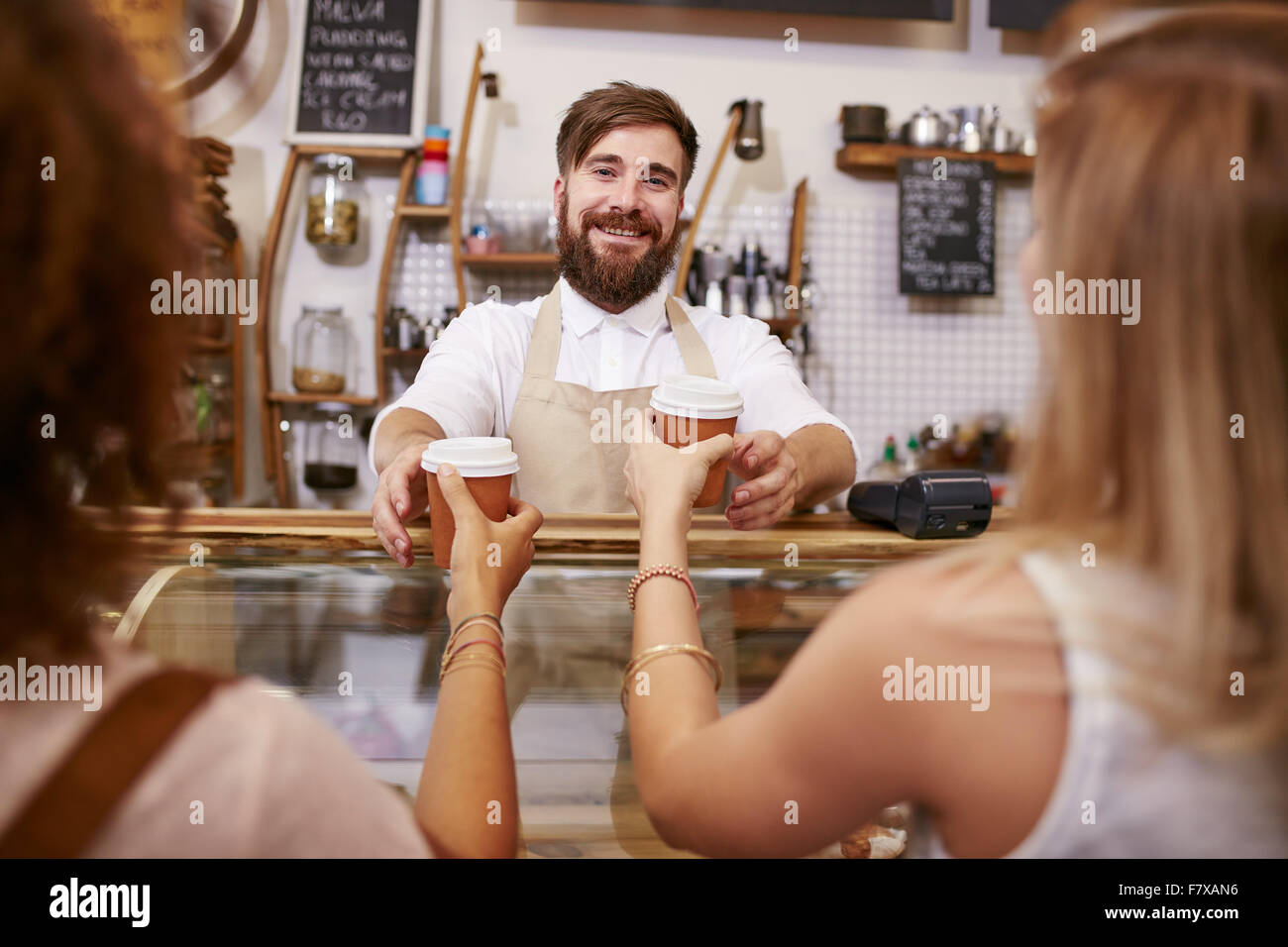 Friendly young man with beard standing behind the cafe counter and serving coffee for two women. Young man working in restaurant Stock Photo