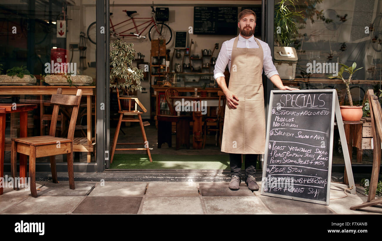 Portrait of young man standing at the entrance of his coffee shop with a black board. Barista standing in the doorway of a resta Stock Photo