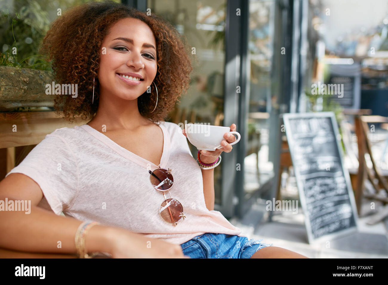 Portrait of young woman drinking coffee. African woman sitting at cafe holding a cup of coffee, looking at camera smiling. Stock Photo