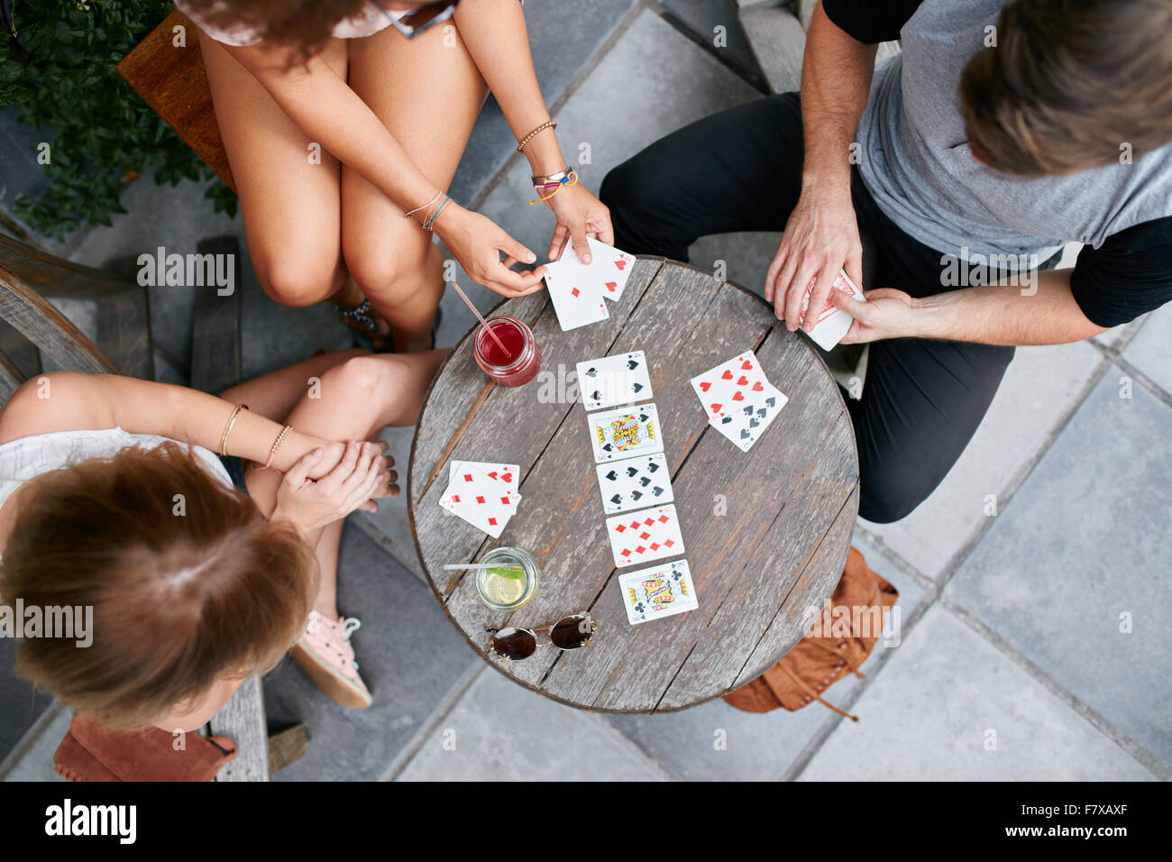 Top view of three young people playing cards at sidewalk cafe. Young people sitting around a coffee table and playing card game. Stock Photo