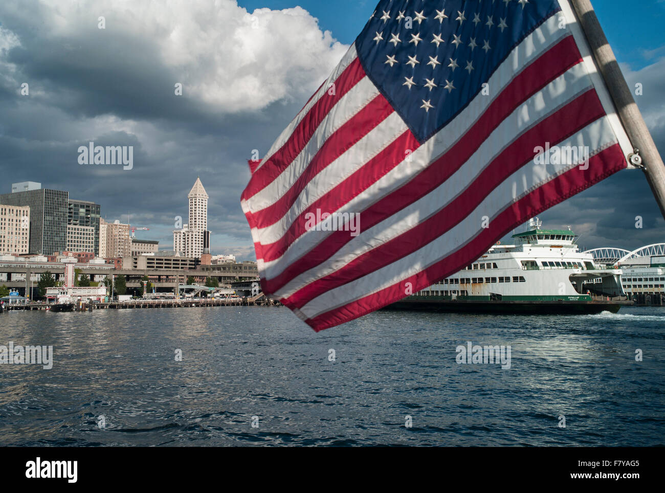 United States, Washington, Seattle, Flag of the United States on the stern of a boat leaving the waterfront with a ferryboat Stock Photo