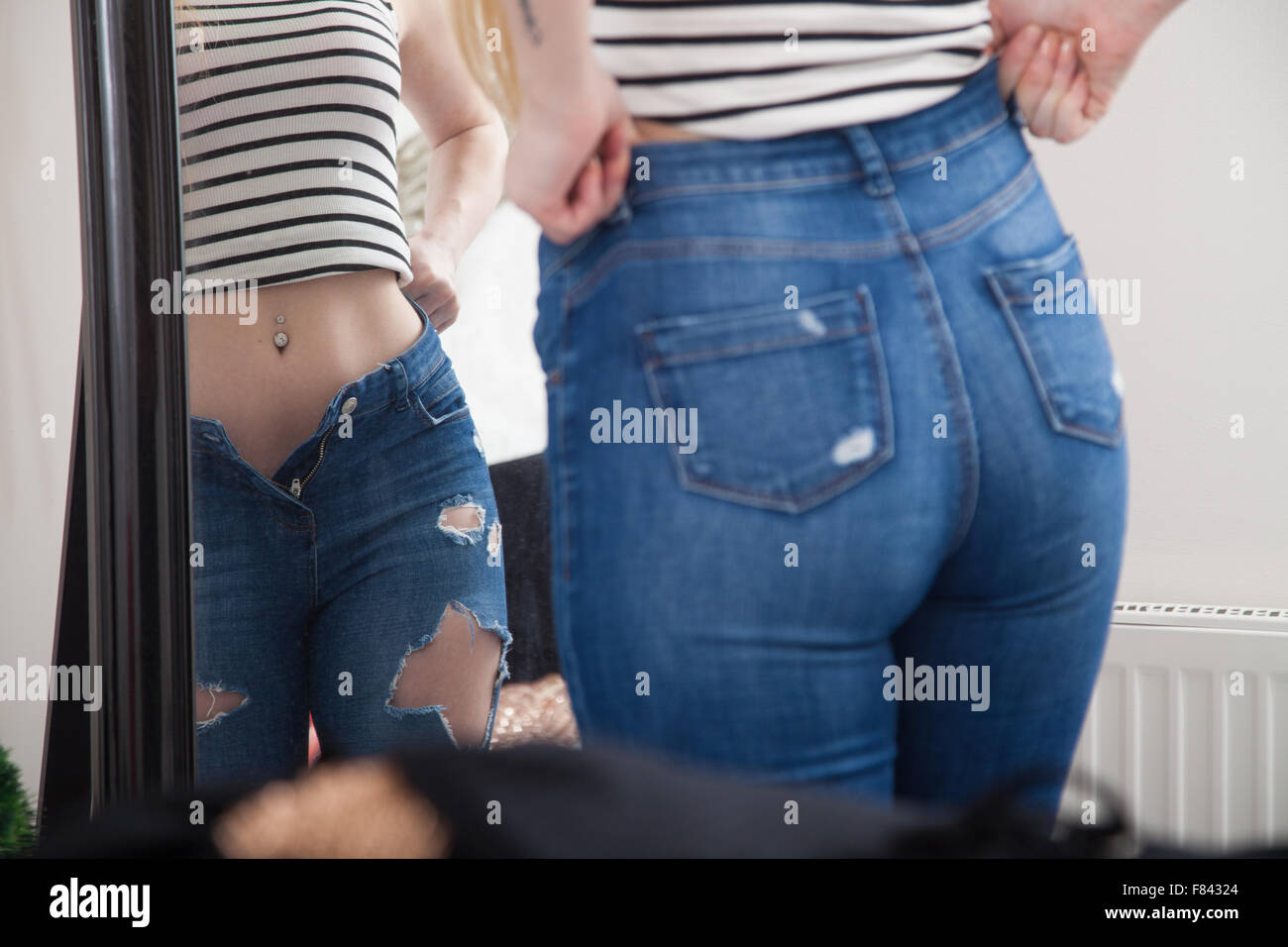A woman at home pulling on tight jeans in front of a bedroom mirror. Stock Photo