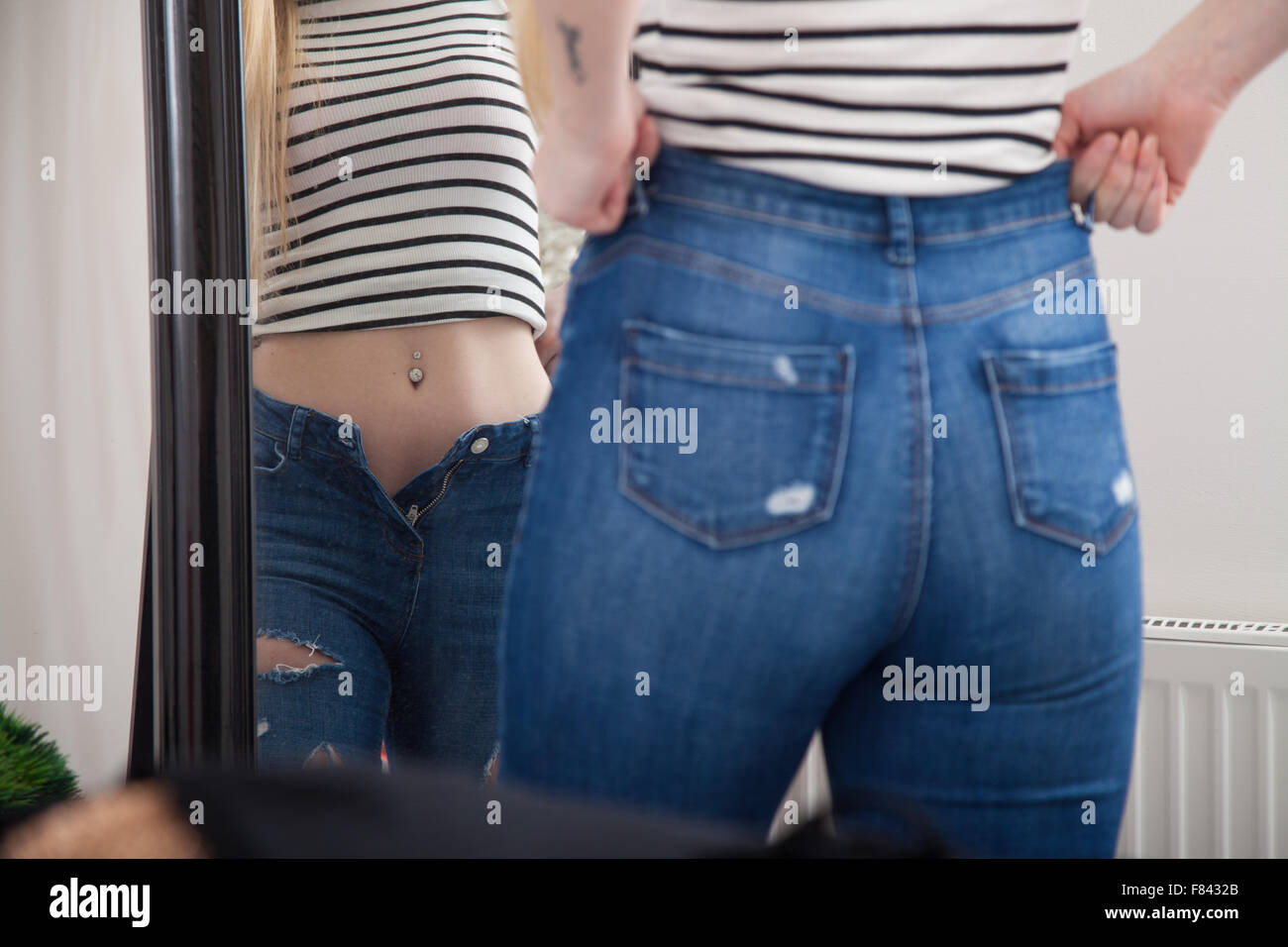 A woman at home pulling on tight jeans in front of a bedroom mirror. Stock Photo