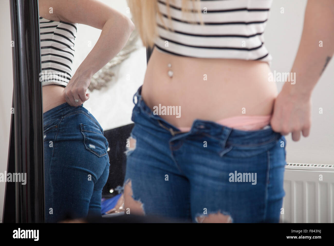A woman at home pulling on tight jeans in front of a bedroom mirror. Stock Photo