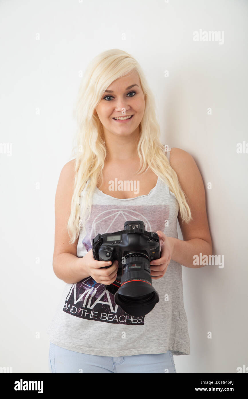 Pretty blond woman leaning against an inside wall holding a DSLR camera. Stock Photo
