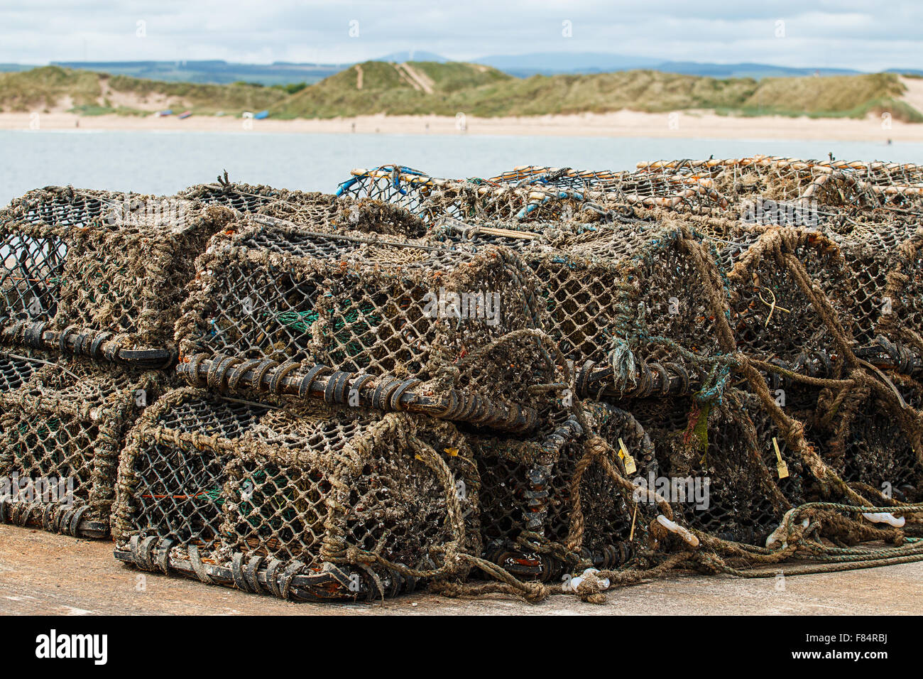 Lobster pots stacked in pile on a harbour with a sandy bay in the background Stock Photo