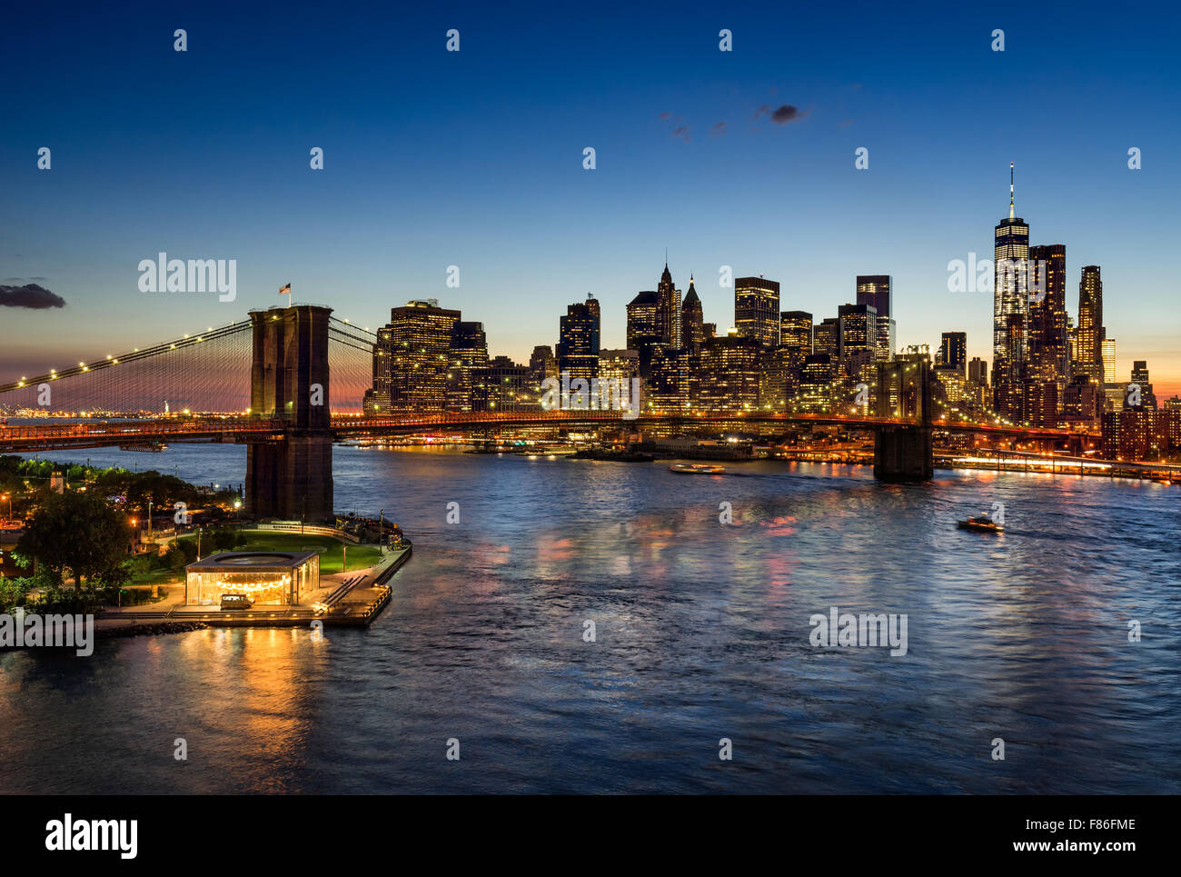 Brooklyn Bridge and illuminated Lower Manhattan at twilight. Financial District skyscrapers reflect in the East River, New York. Stock Photo