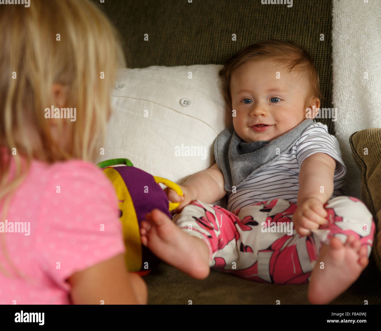 Happy baby leaning back in a sofa and watching older sister Stock Photo