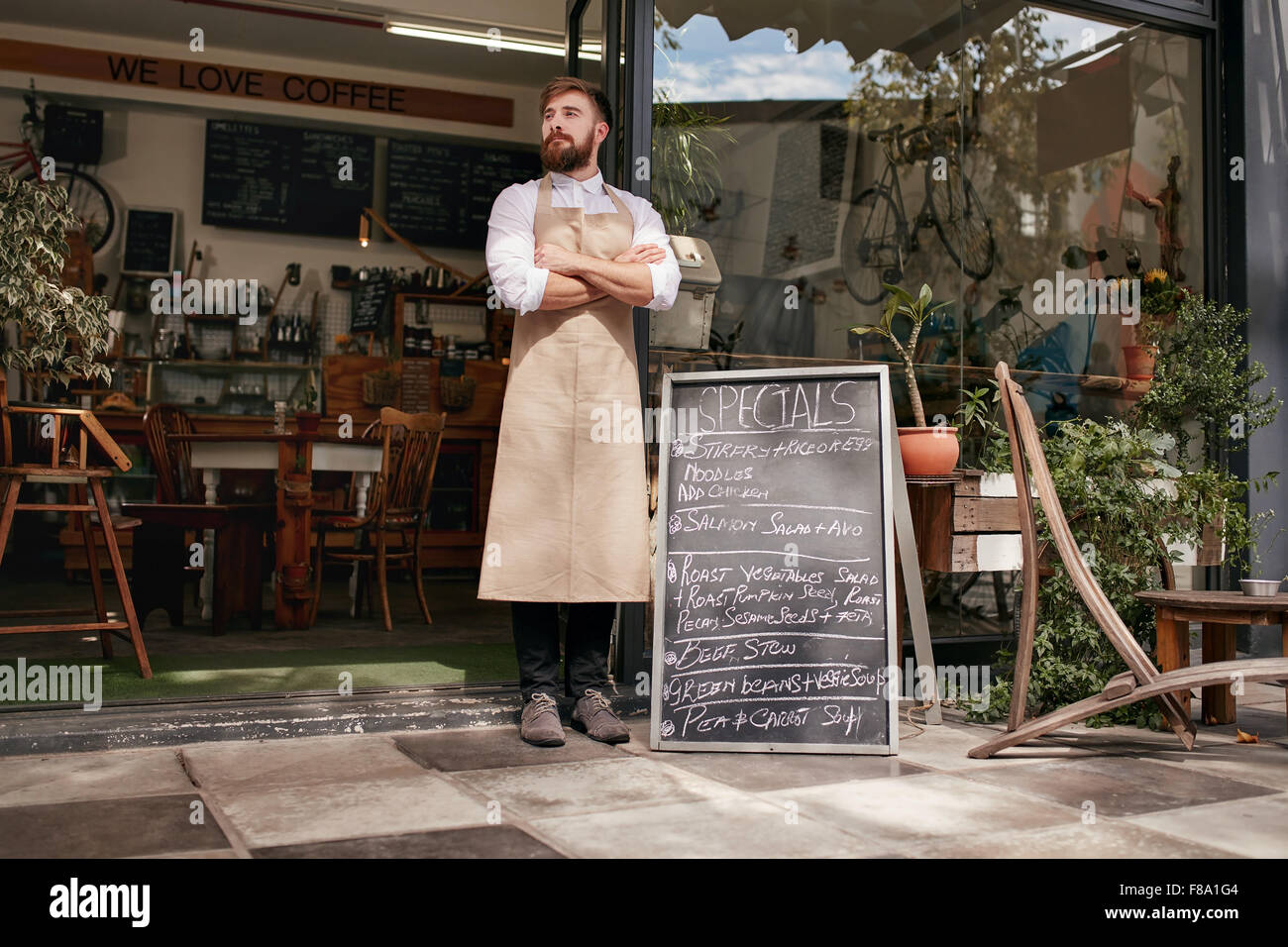Full length shoot of a young waiter standing in door of a cafe. Young man with beard wearing an apron  standing with her arms cr Stock Photo
