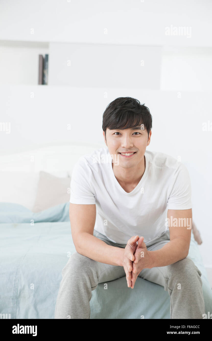 Smiling man sitting on the corner of the bed with hands folded and resting his elbows on his knees Stock Photo