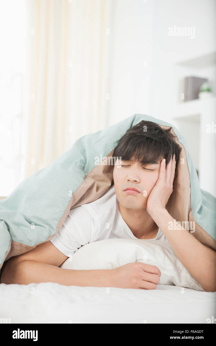 Tired man covered with a comforter lying on his stomach on bed with eyes closed Stock Photo