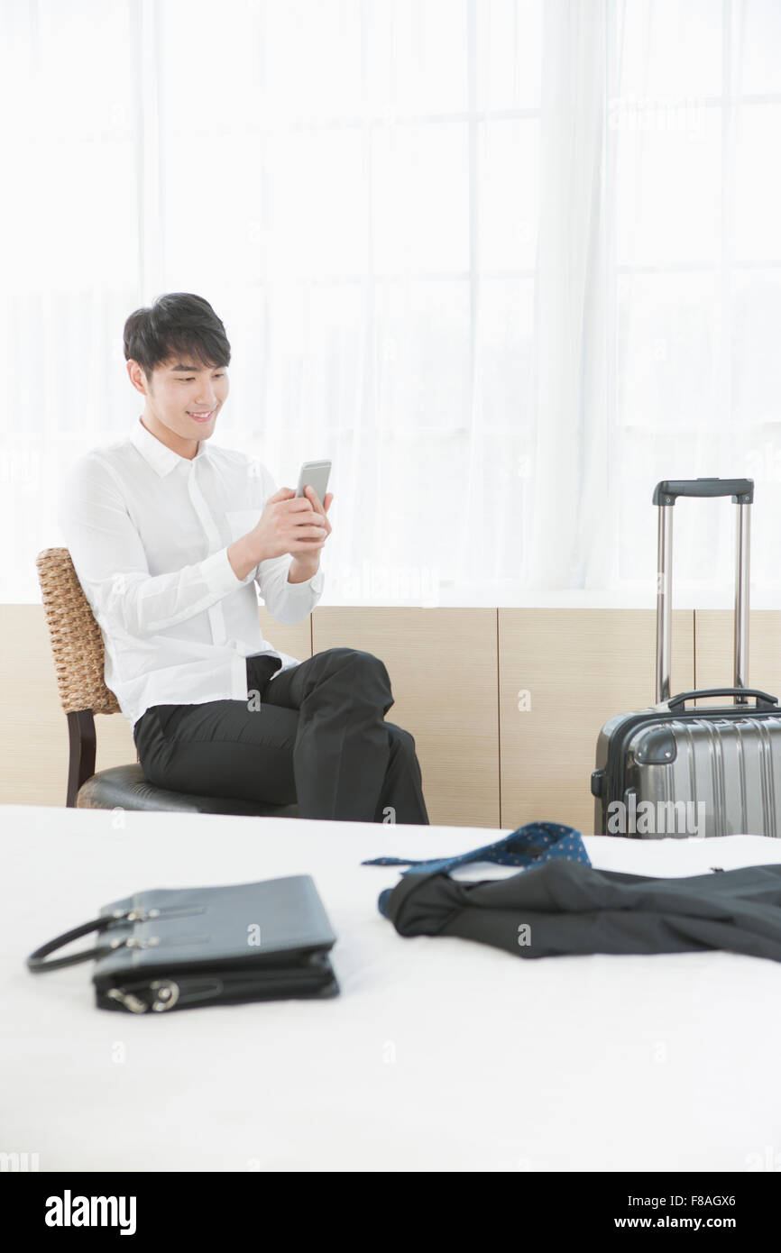 Man sitting on the chair and looking at his phone next to his suitcase and behind the bed Stock Photo