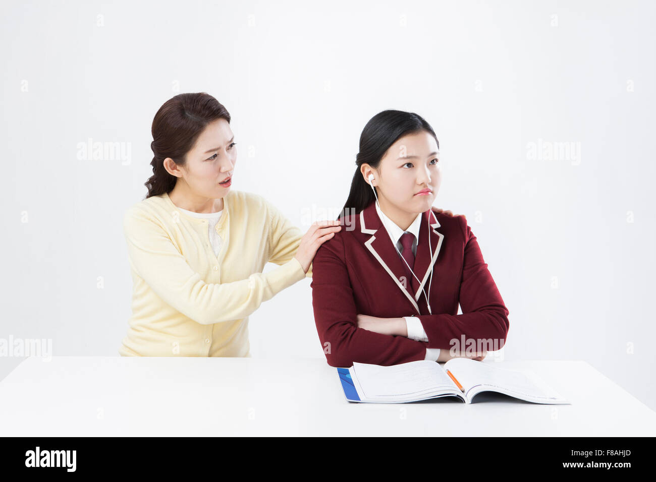 Mother in dissatisfaction touching her daughter's shoulder and daughter listening to music at desk with an open book Stock Photo