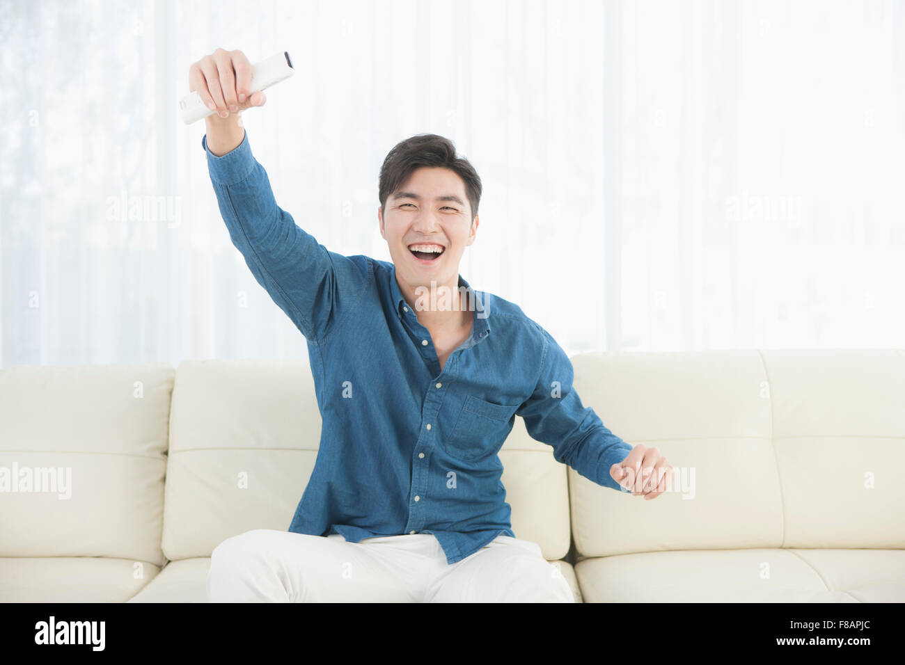 Smiling excited young man cheering holding hand with remote control sitting on sofa Stock Photo