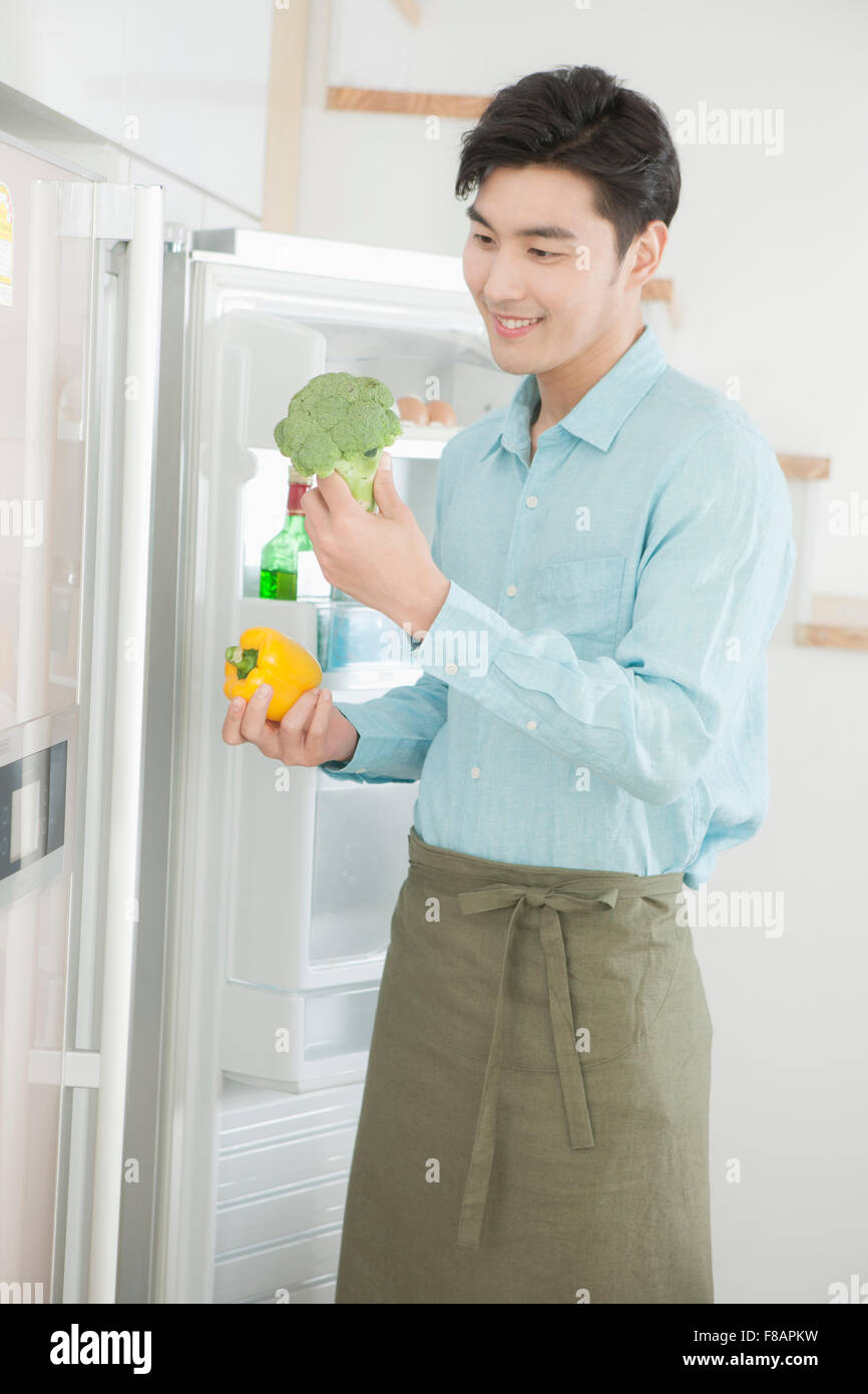 Smiling young man in apron holding paprika and broccoli before open refrigerator Stock Photo