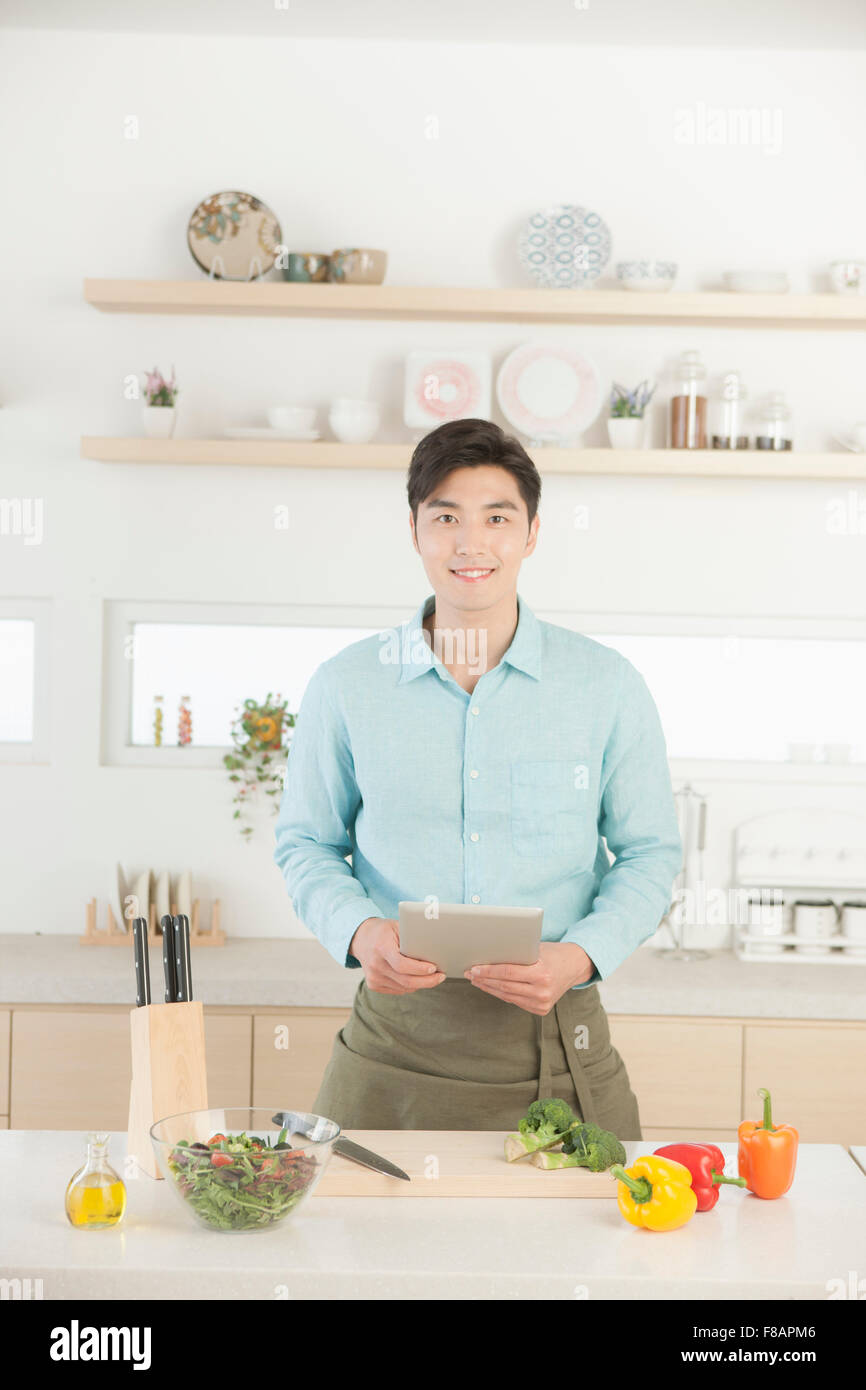 Smiling young man in apron holding tablet staring at front in the kitchen Stock Photo