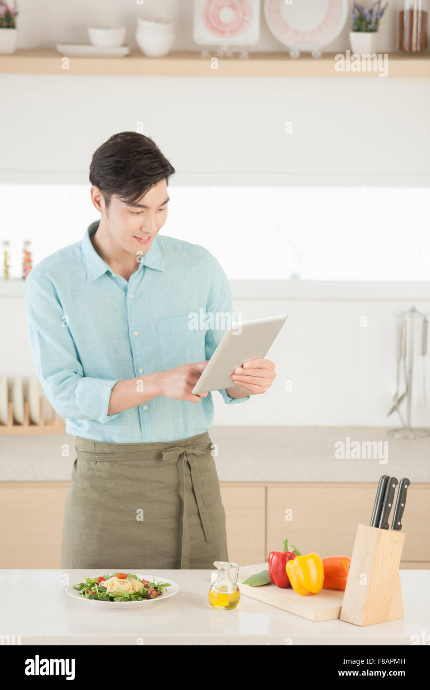 Smiling young man in apron holding and looking down at a tablet in the kitchen Stock Photo