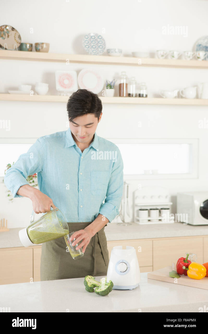 Smiling young man in apron pouring vegetable juice into a glass looking down in the kitchen Stock Photo